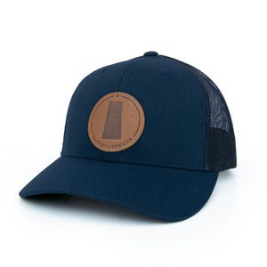 May include: A navy blue trucker hat with a brown leather patch featuring the outline of the province of Saskatchewan and the word "Saskatchewan".
