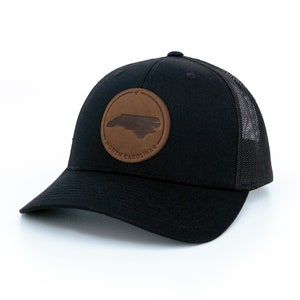 May include: Black trucker hat with a brown leather patch featuring the outline of North Carolina and the text "NORTH CAROLINA".
