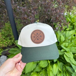 May include: A baseball cap with an off-white crown and a dark green brim. Centered on the front is a circular brown patch featuring a cross design with the letters A, C, and N, along with a small maple leaf icon at the bottom center.