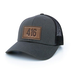 May include: A dark grey trucker hat with a black mesh back. The hat has a brown leather patch with the number "416" stitched in brown thread.