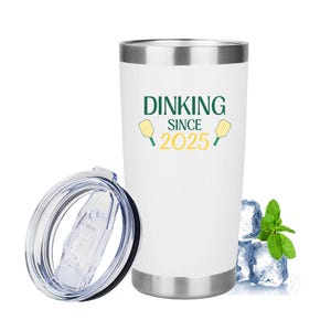 May include: White stainless steel tumbler with a silver rim and base. The tumbler has the text "DINKING SINCE 2025" in green and yellow. The lid is off to the side. Ice cubes and mint sprigs are next to the tumbler.