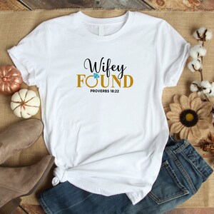 May include: White t-shirt with the text "Wifey Found" in gold lettering with a diamond graphic above the word "Found". The text "Proverbs 18:22" is printed below the main text.