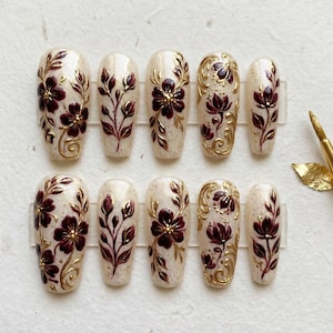 May include: A set of ten artificial nails with a floral design. The nails are a light beige color with a burgundy floral pattern and gold accents. The nails are arranged in two rows of five, with a gold-tipped brush in the background.