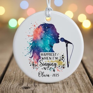 May include: A round, white ceramic ornament with a watercolor design. It features a silhouette of a person singing into a microphone, with the text "Happiest When I'm Singing" and the name "Olivia - 2025". The design includes musical notes and colorful splashes.