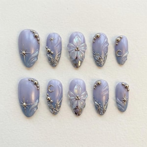 Pode incluir: Um conjunto de dez unhas postiças ovais de cor lavanda. Cada unha apresenta enfeites prateados, incluindo estrelas, luas, desenhos florais e pequenas contas metálicas. As unhas têm um acabamento brilhante.