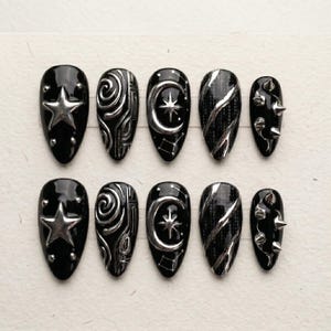 K&ouml;nnte beinhalten: Ein Set aus zehn mandelf&ouml;rmigen Press-on-N&auml;geln in gl&auml;nzendem Schwarz mit silbernen 3D-Verzierungen. Die Designs umfassen einen gro&szlig;en Stern, wirbelnde Metallmuster, einen Halbmond mit Stern, diagonale silberne Linien und kleine silberne Nieten.