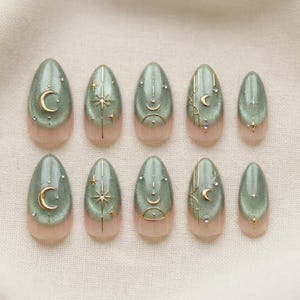 Peut inclure: Dix faux ongles ovales. Chaque ongle présente une base chatoyante vert sauge avec une zone de cuticule couleur chair. Des motifs célestes dorés incluent des lunes, des étoiles et des lignes. Certains ongles ont de petits strass transparents.