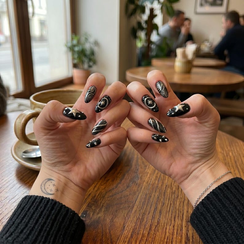 K&ouml;nnte beinhalten: Manik&uuml;rte N&auml;gel mit schwarzer Basis und silbernen Metallic-Akzenten, darunter Stern-Charms, wirbelnde Linienmuster und kleine Nieten. Das Design befindet sich auf mandelf&ouml;rmigen N&auml;geln, und am Handgelenk ist ein kleiner Mondsichel-Tattoo sichtbar.