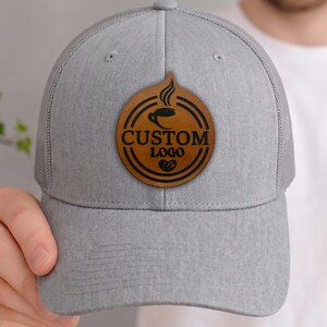 May include: A gray trucker hat with a mesh back and a curved brim. The hat features a circular brown leather patch with the words "CUSTOM LOGO" and a coffee cup design. The hat is being held by a person.