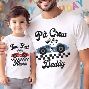 May include: Two white t-shirts with racing-themed graphics. The smaller shirt features a red race car, checkered flags, and the text Two Fast Austin. The larger shirt features a blue race car, checkered flags, and the text Pit Crew Daddy.