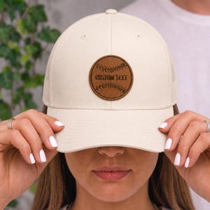 May include: Cream-colored baseball cap with a brown leather patch featuring the text "CUSTOM TEXT" and baseball stitching. The cap has a mesh back and a curved brim. A stylish accessory for sports enthusiasts.