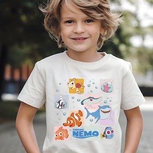 May include: A white t-shirt featuring colorful cartoon characters from the movie "Finding Nemo." The design includes images of fish, a turtle, and a shark, with the movie title in blue. Bubbles are scattered around the characters.