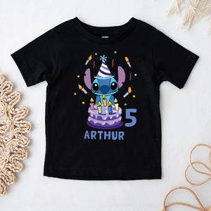 May include: Black t-shirt featuring a cartoon character with a birthday cake and candles. The design includes the number "5" and the name "ARTHUR" in blue. The character is wearing a party hat.