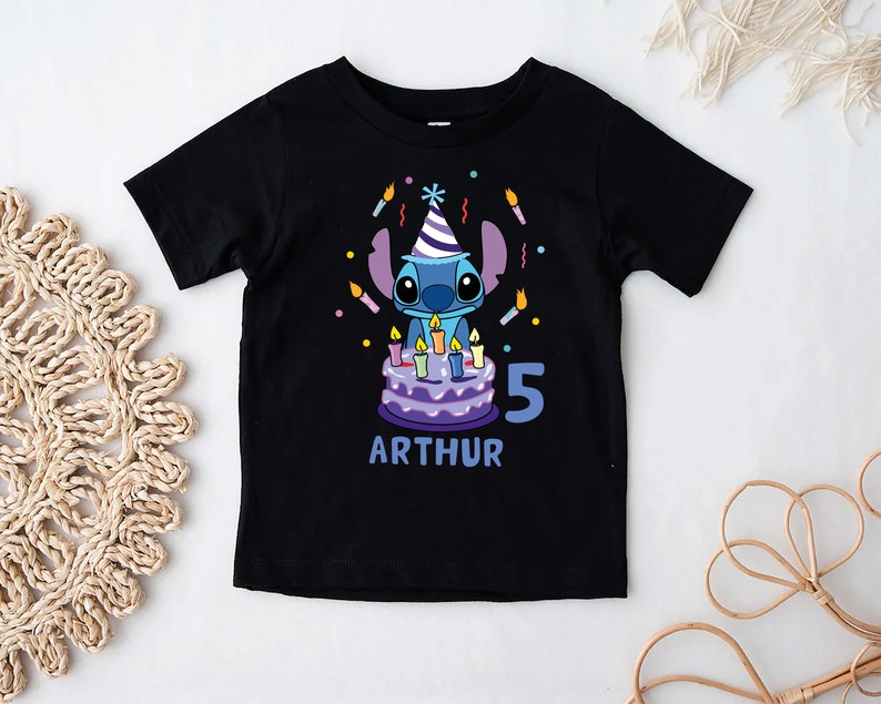 May include: Black t-shirt featuring a cartoon character with a birthday cake and candles. The design includes the number "5" and the name "ARTHUR" in blue. The character is wearing a party hat.