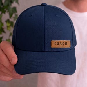 Könnte beinhalten: Marineblaue Baseballkappe mit Mesh-Rückseite und gebogenem Schirm. Ein rechteckiger, hellbrauner Lederaufnäher auf der Vorderseite zeigt das Wort "COACH" über "YOUR NAME". Die Kappe hat ein klassisches, sportliches Design.
