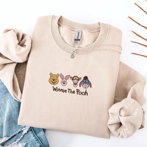 May include: A beige crewneck sweatshirt featuring an embroidered graphic of Winnie the Pooh, Piglet, Tigger, and Eeyore faces arranged in a row. Below the characters, the text Winnie The Pooh is embroidered in a dark brown, rounded script font.