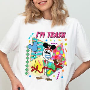 May include: White t-shirt with a colourful graphic featuring the text "I'M TRASH" in pink letters. The graphic includes a cartoon character with a spoon-shaped head, Mickey Mouse ears, and a blue balloon.