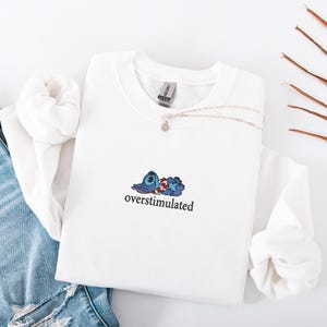May include: A white crewneck sweatshirt featuring an embroidered graphic of a blue alien character lying down while wearing a red and white floral lei. The text "overstimulated" is embroidered in black lowercase letters below the character.