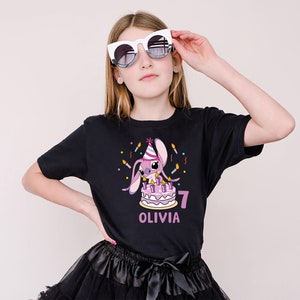 May include: A black t-shirt featuring a cartoon bunny with a birthday cake, candles, and the number 7. The name "OLIVIA" is printed below the number. The person is wearing white sunglasses and a black skirt.