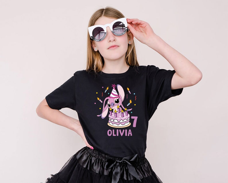 May include: A black t-shirt featuring a cartoon bunny with a birthday cake, candles, and the number 7. The name "OLIVIA" is printed below the number. The person is wearing white sunglasses and a black skirt.