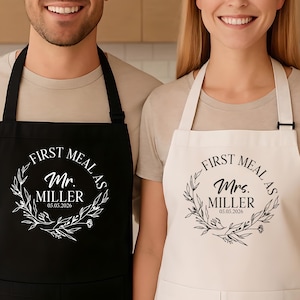 May include: Matching black and white aprons with the text "First Meal As Mr. & Mrs. Miller 05.05.2026" in a decorative wreath design. The aprons are worn by a couple, suggesting a wedding or anniversary theme.