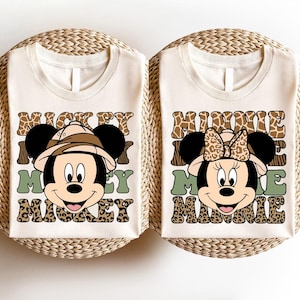 May include: Two white t-shirts with a safari-themed Mickey Mouse and Minnie Mouse design. The shirts feature a brown, green, and white pattern with the text "Mickey Mouse" and "Minnie Mouse".