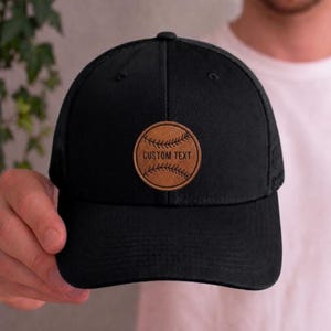 May include: Black baseball cap with a brown leather patch. The patch features a baseball design with the words "CUSTOM TEXT" in white. The cap has a curved brim and a mesh back.