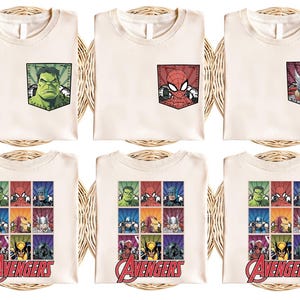 May include: Cream-colored t-shirts featuring Marvel superheroes. Some shirts have pocket designs with Hulk, Spider-Man, and Thor. Other shirts display a grid of Avengers characters with the word "Avengers" in red.