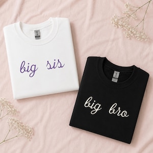 Może przedstawiać: Biała bluza z okrągłym dekoltem z fioletowym napisem big sis oraz czarna bluza z białym napisem big bro. Obie części garderoby mają długie rękawy, ściągacze przy dekolcie oraz prostą czcionkę imitującą pismo odręczne dla nadruku.