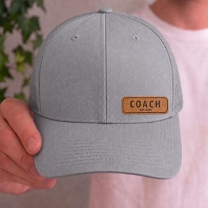 Könnte beinhalten: Graue Baseballkappe mit Mesh-Rückseite und gebogenem Schirm. Die Kappe hat einen braunen Lederaufnäher mit dem Wort "COACH" und dem Text "YOUR NAME" aufgedruckt. Die Kappe wird von einer Person hochgehalten.