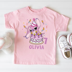 May include: A light pink t-shirt featuring a cartoon character with a birthday cake and candles. The shirt has the name "Olivia" and the number "7" printed on it. White sneakers are next to the shirt.