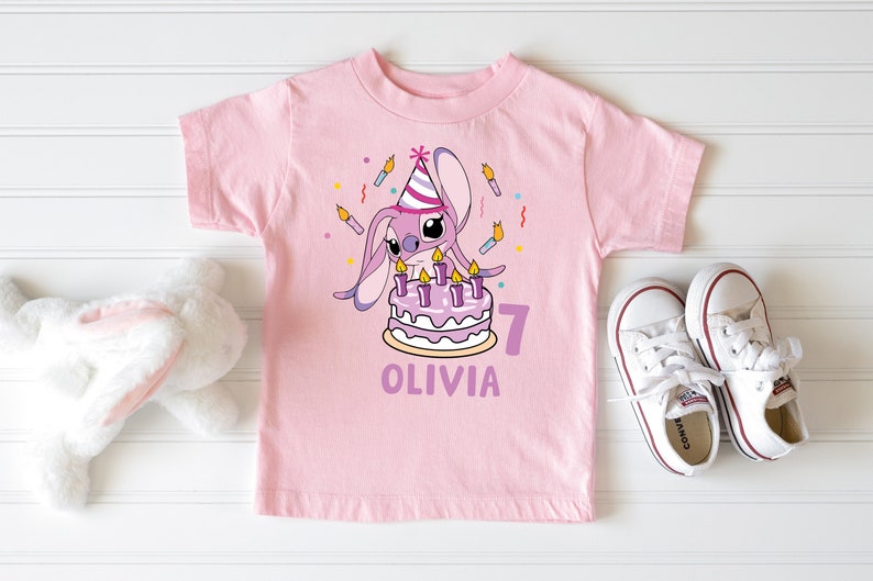 May include: A light pink t-shirt featuring a cartoon character with a birthday cake and candles. The shirt has the name "Olivia" and the number "7" printed on it. White sneakers are next to the shirt.