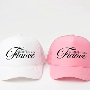 May include: Two trucker caps, one white and one pink. The white cap has the text "Boyfriend Fiance" in black script, and the pink cap says "Girlfriend Fiance" in black script. Both caps have a mesh back.