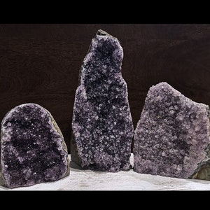 May include: Four amethyst crystal clusters, each with a unique shape and size, are displayed on a white surface. The crystals are a deep purple color and have a natural, rough texture.