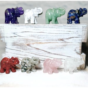 May include: Seven small elephant figurines carved from different coloured stones. The elephants are arranged in a row on a white wooden surface. The stones are amethyst, white, green, blue, red, green, and white.