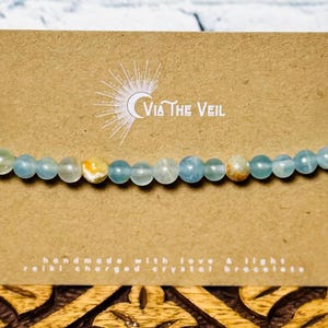 Puede incluir: Un brazalete de cristal azul claro con algunas cuentas amarillas y marrones. El brazalete est&aacute; sobre una tarjeta marr&oacute;n con el texto "Via The Veil" y "hecho a mano con amor y luz, brazalete de cristal cargado con Reiki".