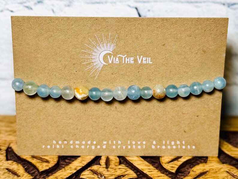 Puede incluir: Un brazalete de cristal azul claro con algunas cuentas amarillas y marrones. El brazalete est&aacute; sobre una tarjeta marr&oacute;n con el texto "Via The Veil" y "hecho a mano con amor y luz, brazalete de cristal cargado con Reiki".