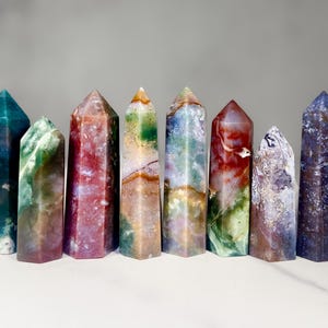 Può includere: Una collezione di sette obelischi di pietre preziose lucide. Gli obelischi presentano una varietà di colori, tra cui sfumature di verde, rosso, viola e blu, con motivi e trame uniche. Ogni obelisco ha una sommità appuntita e una base rettangolare.