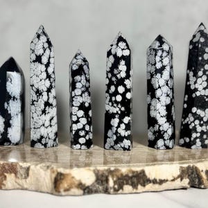 May include: A collection of snowflake obsidian obelisks. These polished stone towers feature a black base color with white snowflake-like patterns. The obelisks vary in height and are displayed on a natural wood slab.