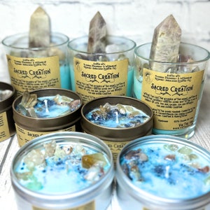 May include: Five teal blue candles with embedded crystals and a clear glass wick. The candles are in various sizes and are labeled "Sacred Creation" with a description of the candle's purpose. The candles are on a white wooden surface.