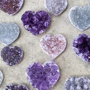 May include: An assortment of heart-shaped amethyst crystals in various shades of purple, lavender, and gray. The crystals have a rough, textured surface and are arranged on a neutral-toned background. These decorative crystals are ideal for home decor.