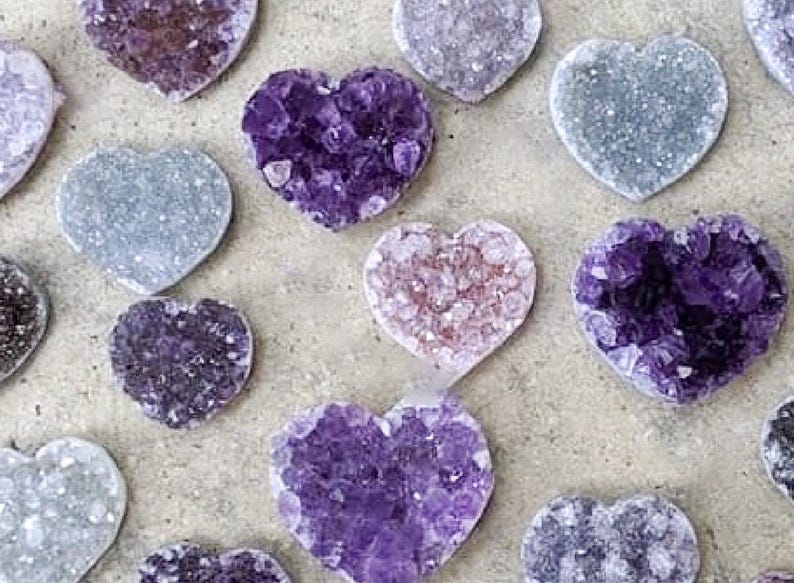 May include: An assortment of heart-shaped amethyst crystals in various shades of purple, lavender, and gray. The crystals have a rough, textured surface and are arranged on a neutral-toned background. These decorative crystals are ideal for home decor.