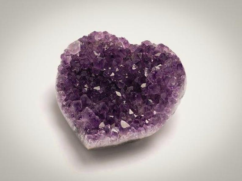May include: A heart-shaped amethyst geode, showcasing a cluster of deep purple crystals. The crystals have a sparkling, crystalline structure, with a light-colored base. The geode is set against a white background.