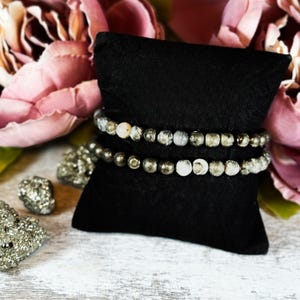 May include: Two beaded bracelets on a black velvet cushion. The bracelets are made of gray and white stones and gold beads.