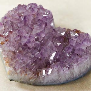 May include: A heart-shaped amethyst geode with a crystalline structure. The purple crystals have a sparkling appearance, with some areas showing a lighter hue. The base of the geode has a white and gray border.