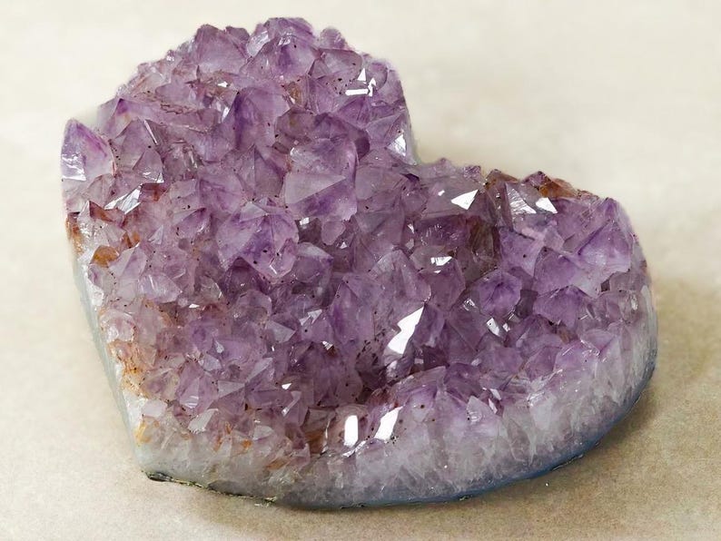 May include: A heart-shaped amethyst geode with a crystalline structure. The purple crystals have a sparkling appearance, with some areas showing a lighter hue. The base of the geode has a white and gray border.