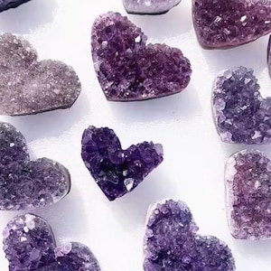 May include: A collection of heart-shaped amethyst crystals in shades of purple are shown from above. The crystals have a rough, textured surface and are arranged on a white background. The crystals vary in size and shape.