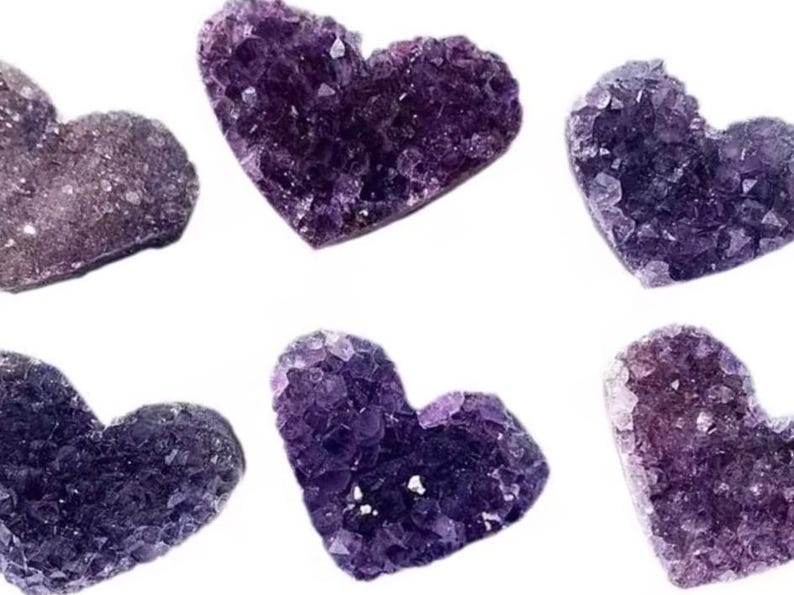 May include: Six heart-shaped amethyst crystal clusters in varying shades of purple and violet. The crystals have a rough, textured surface and are arranged against a white background. Each heart is approximately 2 inches wide.