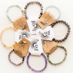 May include: A collection of beaded bracelets in various colors, including green, yellow, purple, and brown. Each bracelet is paired with a tag that reads "VIA THE VEIL CRYSTAL KIDS". The bracelets are arranged on a white surface.