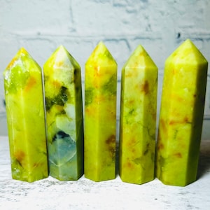May include: Five green, yellow, and white crystal points, also known as wands, are arranged in a row on a white surface. The points are all approximately the same size and shape, and they have a smooth, polished finish.